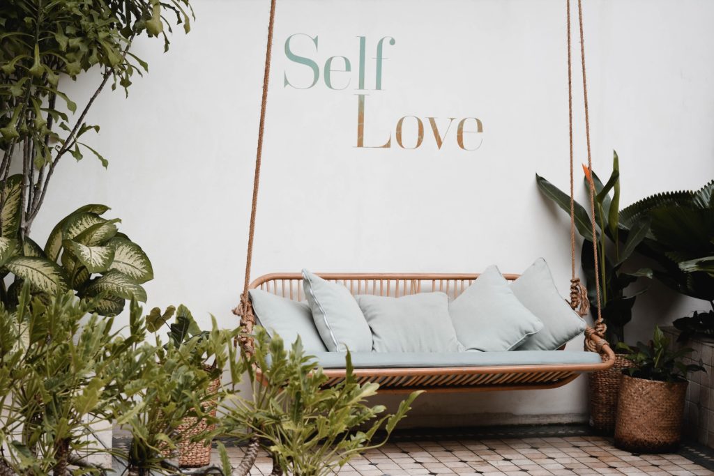 bench swing with pillows and plants surrounding it and the text self love above it