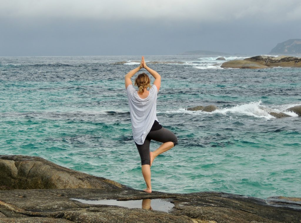 woman standing on rocks in tree pose looking out at ocean waves 