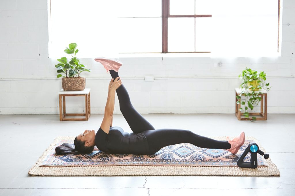 woman in black yoga outfit laying on her back and stretching left leg while holding left ankle with both hands