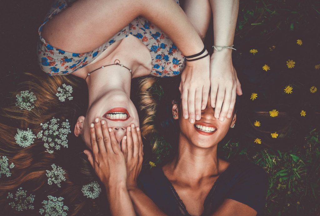 twofemale friends smiling as they lay in a bed of white and yellow flowers with their hands over each other's eyes